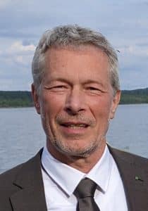 Auf diesem Foto steht der Weniger Stress Experte und Wohnwagenprofessor Stefan Dietz im Anzug und Krawatte lächelnd am Ufer eines Sees und genießt weniger Stress.
